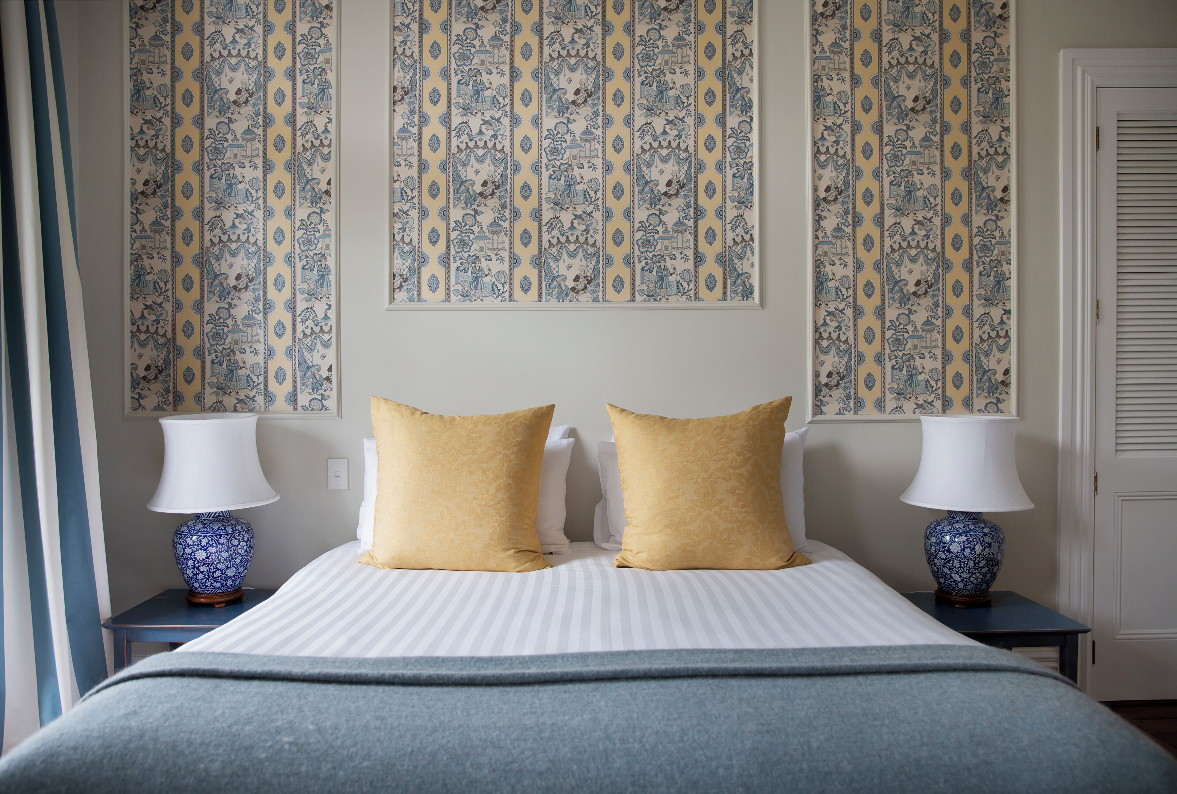 Quarto de hotel com cama, duas mesas de cabeceira, duas luminárias e papel de parede estampado.