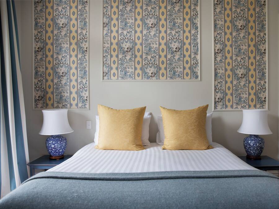 Quarto de hotel com cama, duas mesas de cabeceira, duas luminárias e papel de parede estampado.