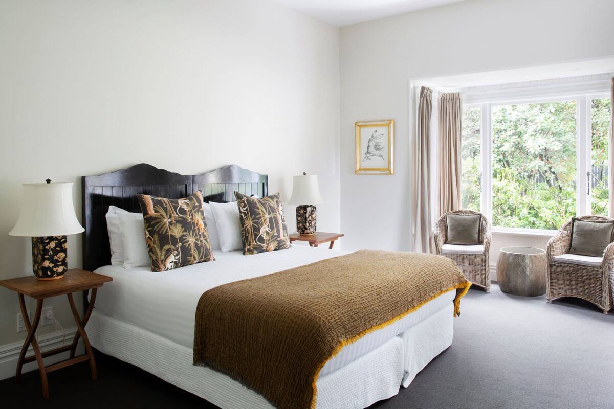 A hotel room with a large bed, two bedside tables with lamps, a wicker chair, a round table, and a window with curtains.