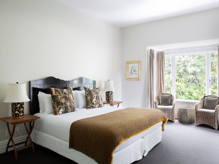 A hotel room with a large bed, two bedside tables with lamps, a wicker chair, a round table, and a window with curtains.