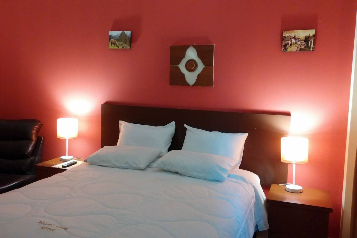 A hotel room with a bed, two chairs, a nightstand with a lamp, and two wall-mounted pictures.