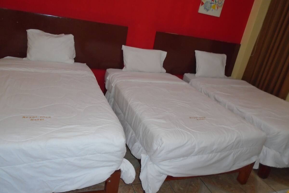 A hotel room with three single beds, each with white bedding and a headboard. The room has a red accent wall and a brown headboard.