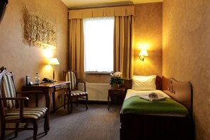 A hotel room with a bed, a desk, a chair, a radiator, and a window with curtains.