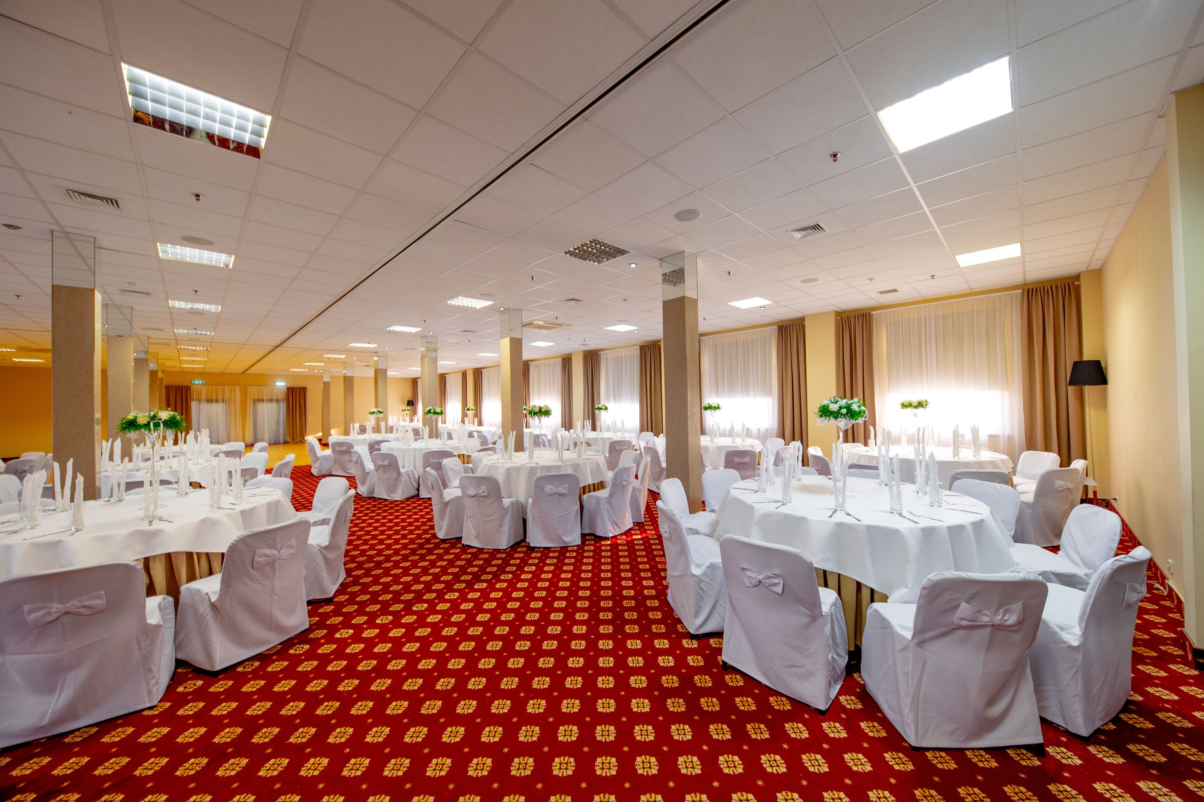 A banquet hall with round tables set for events, featuring a red carpet and white tablecloths.