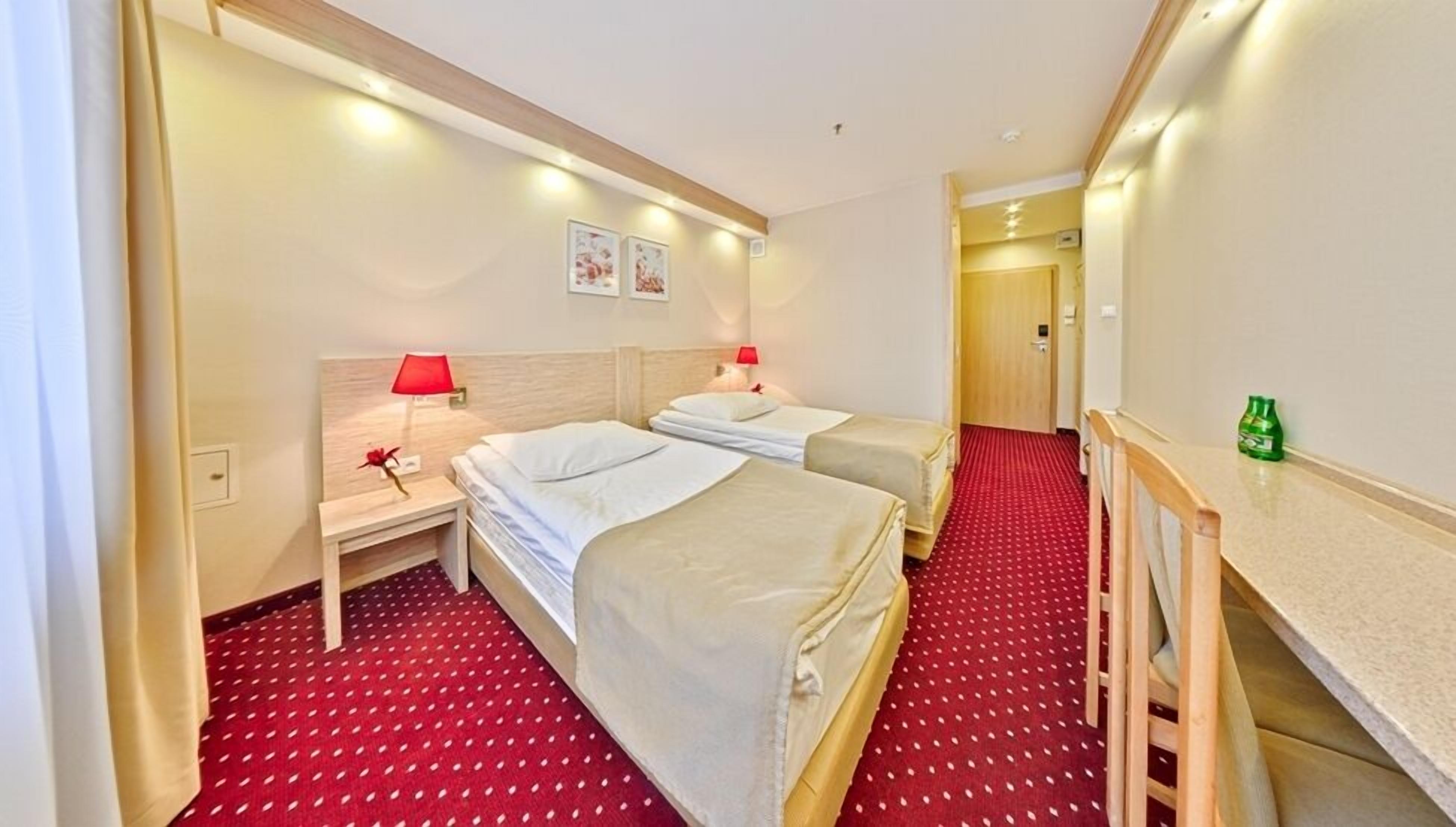 Habitación de hotel con dos camas, un escritorio, una silla, una alfombra roja y una botella verde pequeña sobre el escritorio.