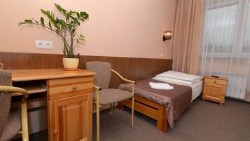 A hotel room with a wooden desk, a chair, a bed with a brown bedspread, a nightstand, a potted plant, and a window with curtains.