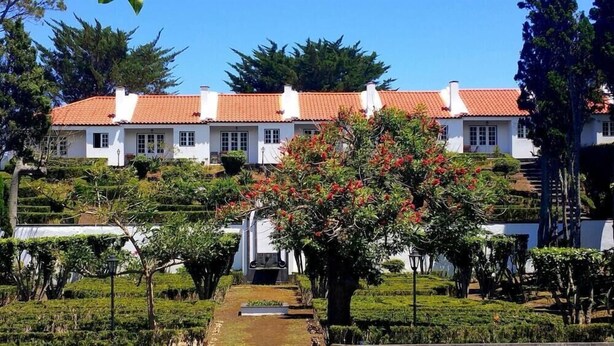Un edificio grande e bianco con tetto in tegole rosse, immerso in un giardino curato con un vialetto centrale che conduce a un piccolo ingresso.