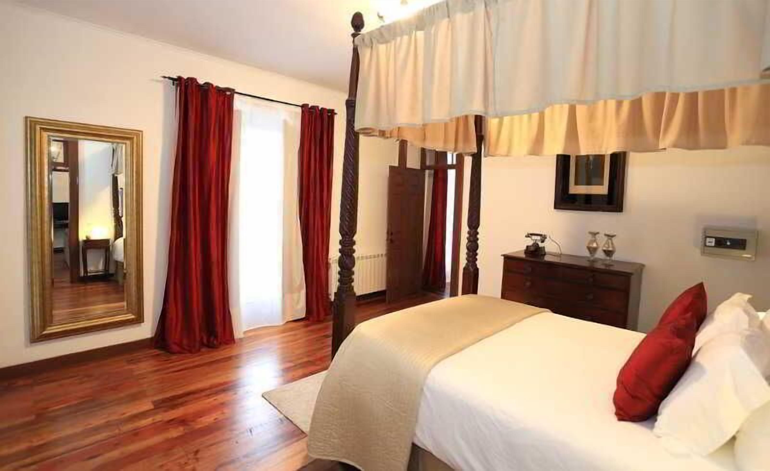 Une chambre à coucher avec un lit à baldaquin, un plancher en bois, une commode, un miroir et des rideaux rouges.