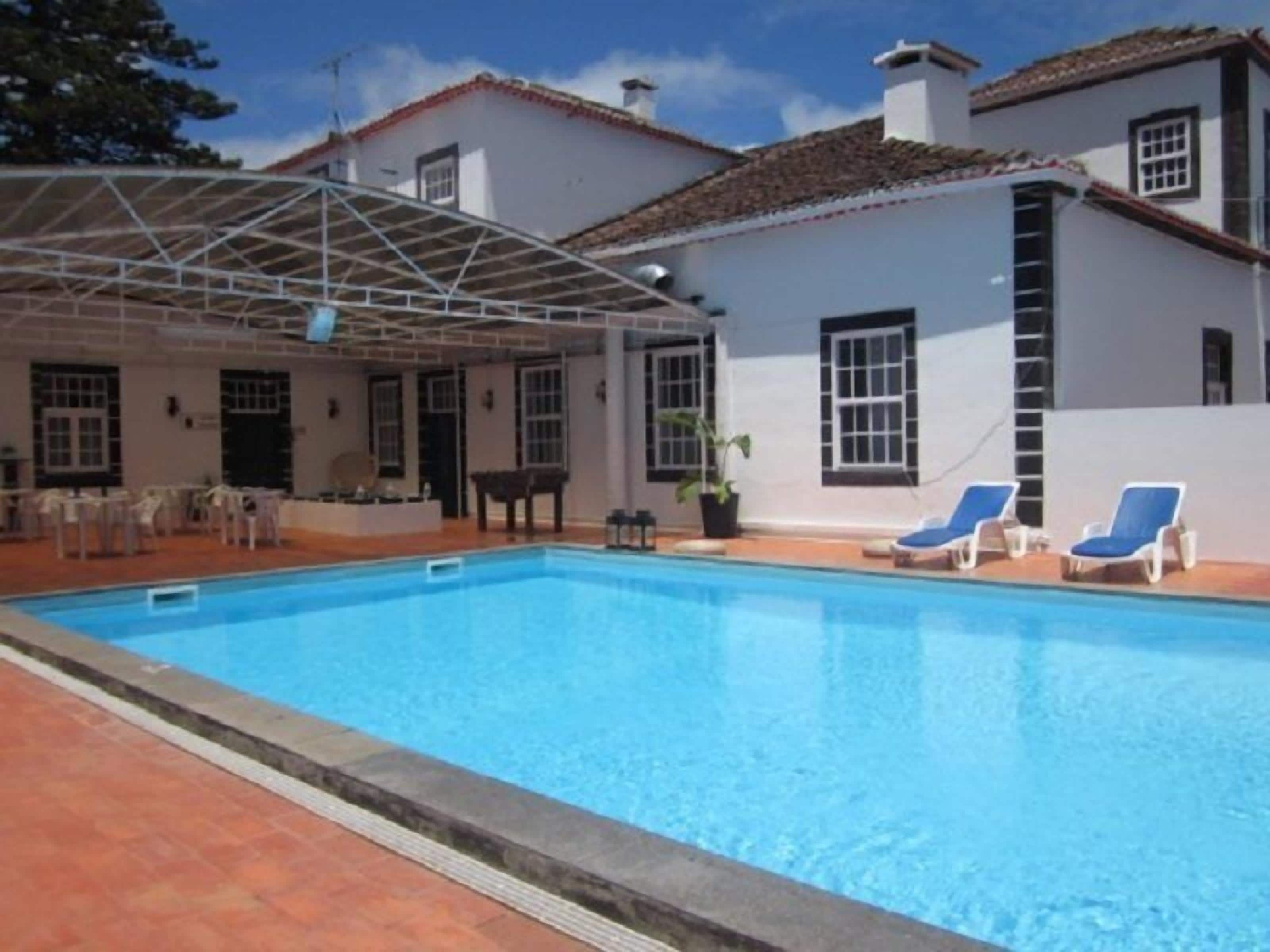 A swimming pool with lounge chairs, a covered patio, and a building with white walls and a tiled roof.