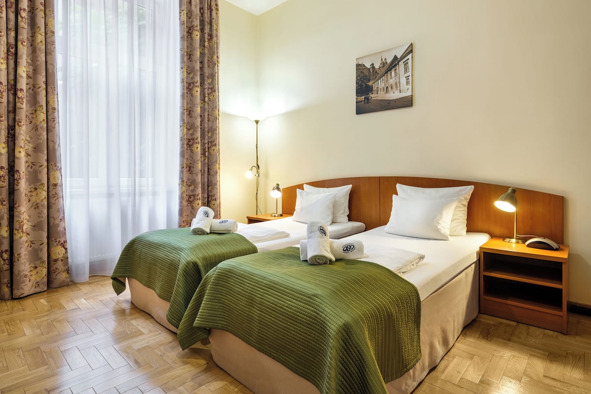 A hotel room with two beds, a wooden headboard, a nightstand with a lamp, a painting on the wall, and a window with floral curtains.