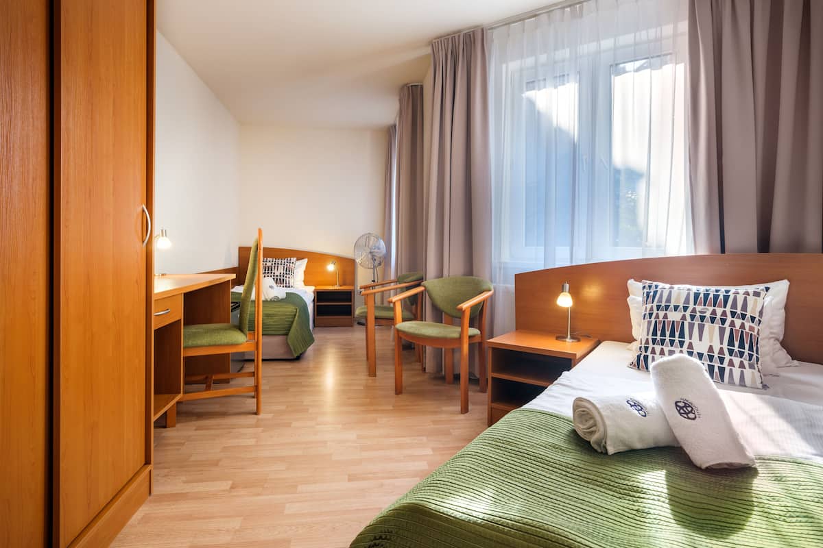 A hotel room with a bed, a desk, two chairs, a bedside table, and a window with curtains.