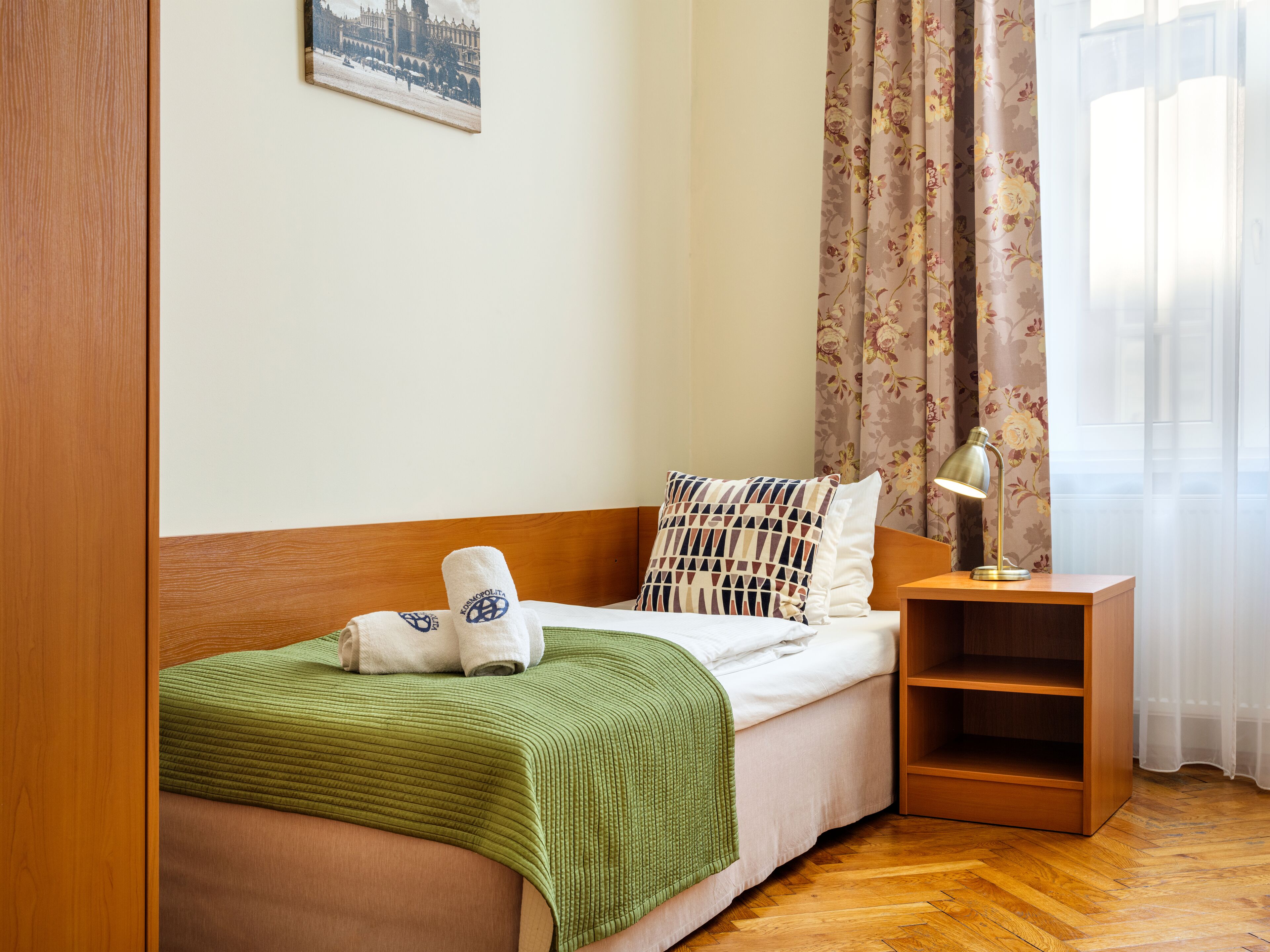 Ein ordentlich bezogenes Bett mit grüner Decke, einem Kopfkissen und zwei Handtüchern mit Logo. Ein Holznachttisch mit Lampe, eine Holzablage und ein gerahmtes Bild an der Wand sind ebenfalls zu sehen.