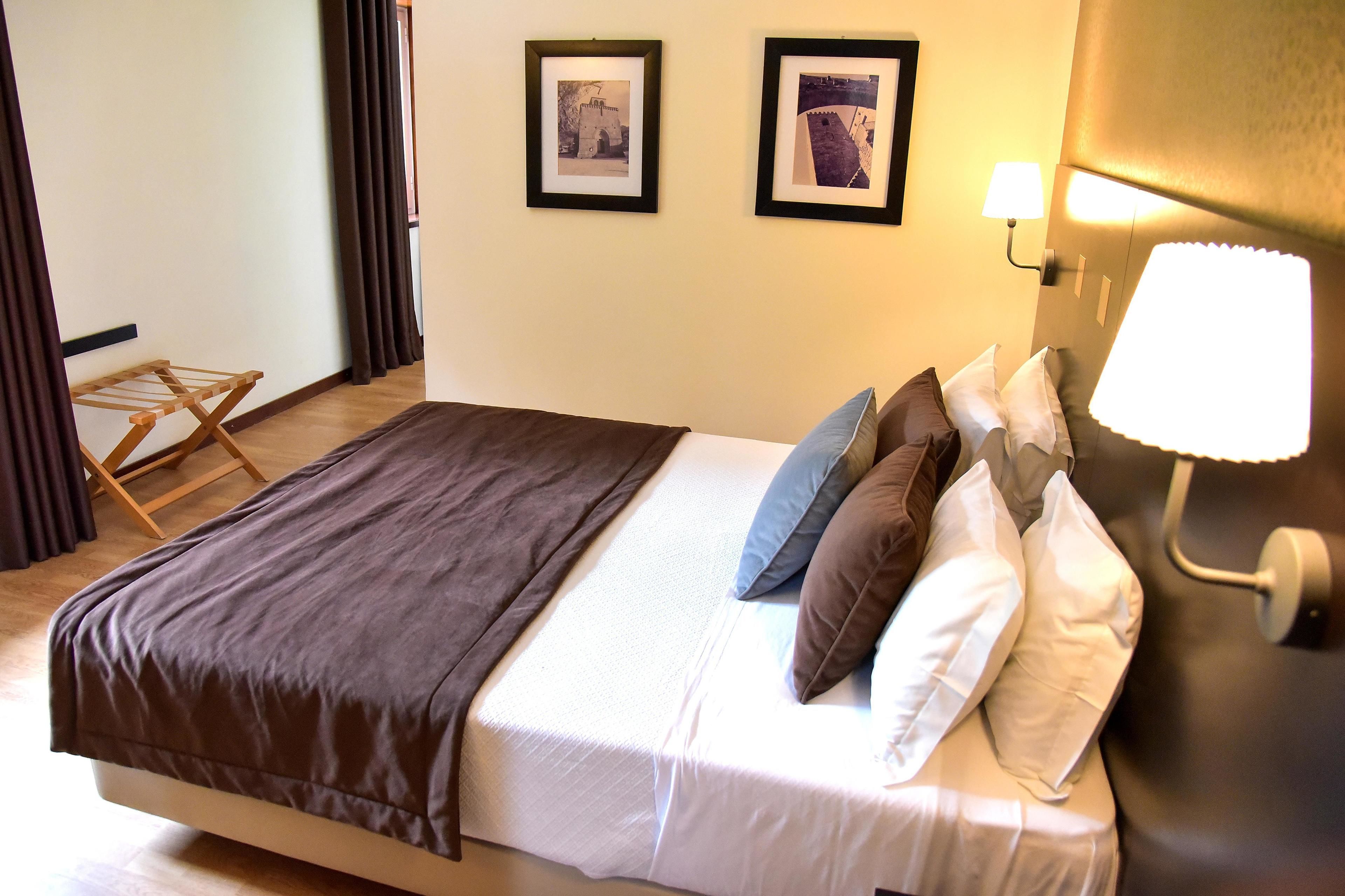 Ein ordentlich bezogenes Bett mit brauner Decke, weißen Leinen und vielen Kissen. Gegen die Wand lehnt ein hölzerner Wäscheständer und es hängen zwei gerahmte Bilder an der Wand.