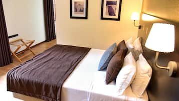 Ein ordentlich bezogenes Bett mit brauner Decke, weißen Leinen und vielen Kissen. Gegen die Wand lehnt ein hölzerner Wäscheständer und es hängen zwei gerahmte Bilder an der Wand.
