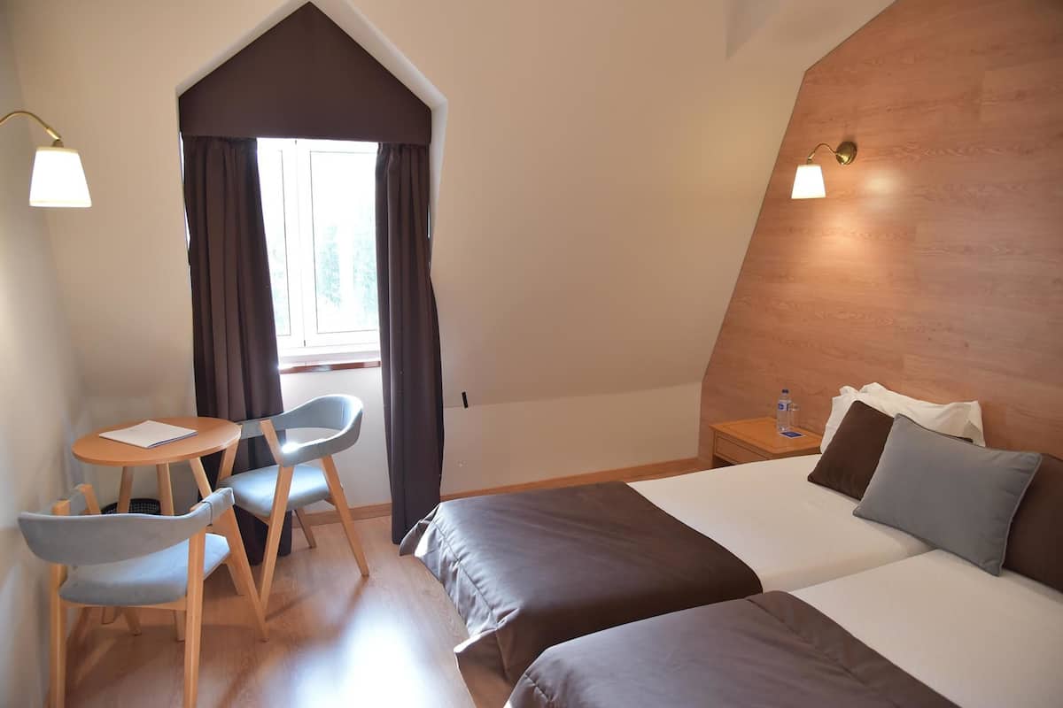 A hotel room with two beds, a small table with chairs, a nightstand with a lamp, and a window with curtains.