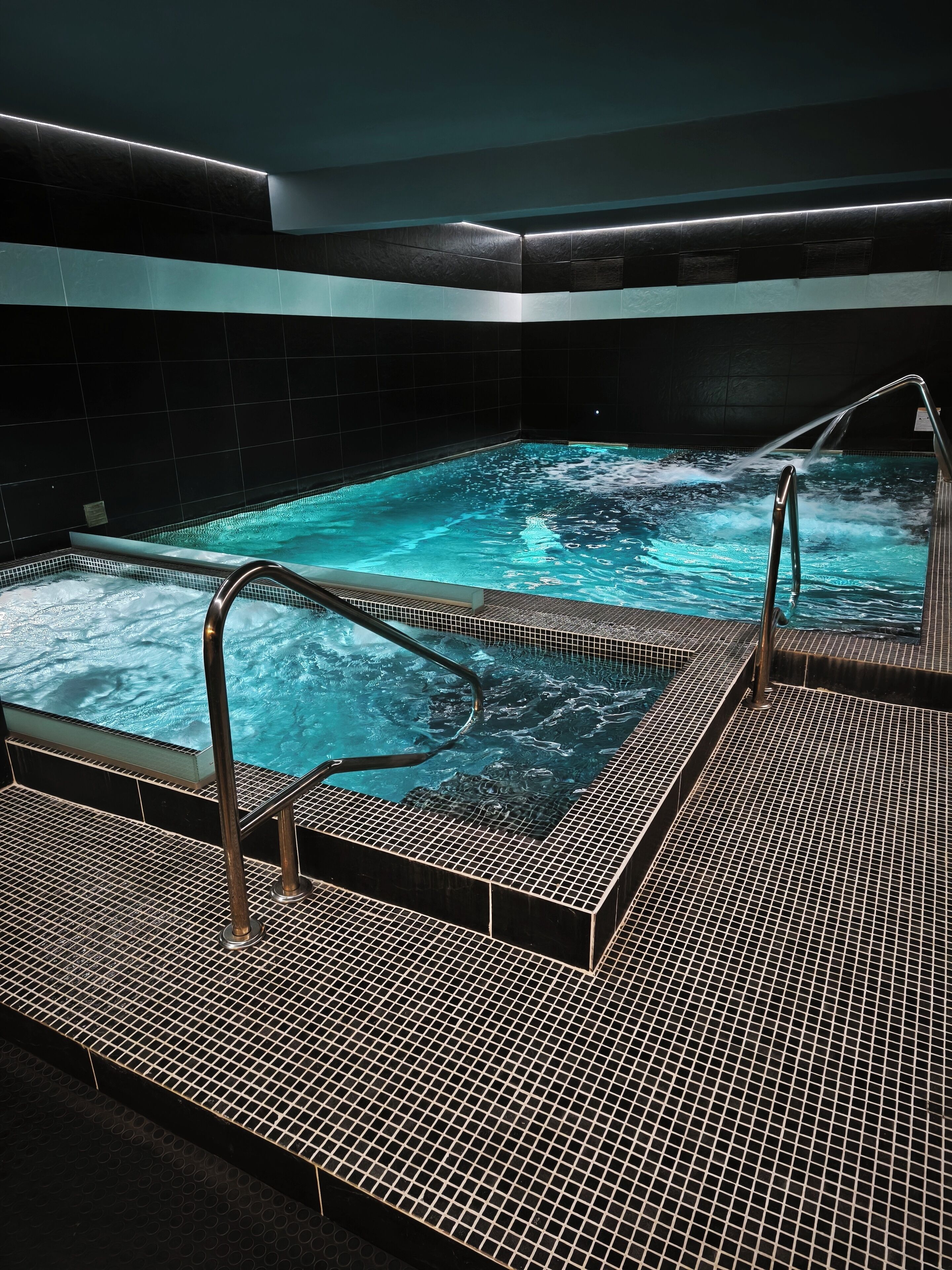 Un área de spa con una bañera de hidromasaje y una piscina, rodeada de paredes cubiertas de baldosas oscuras.