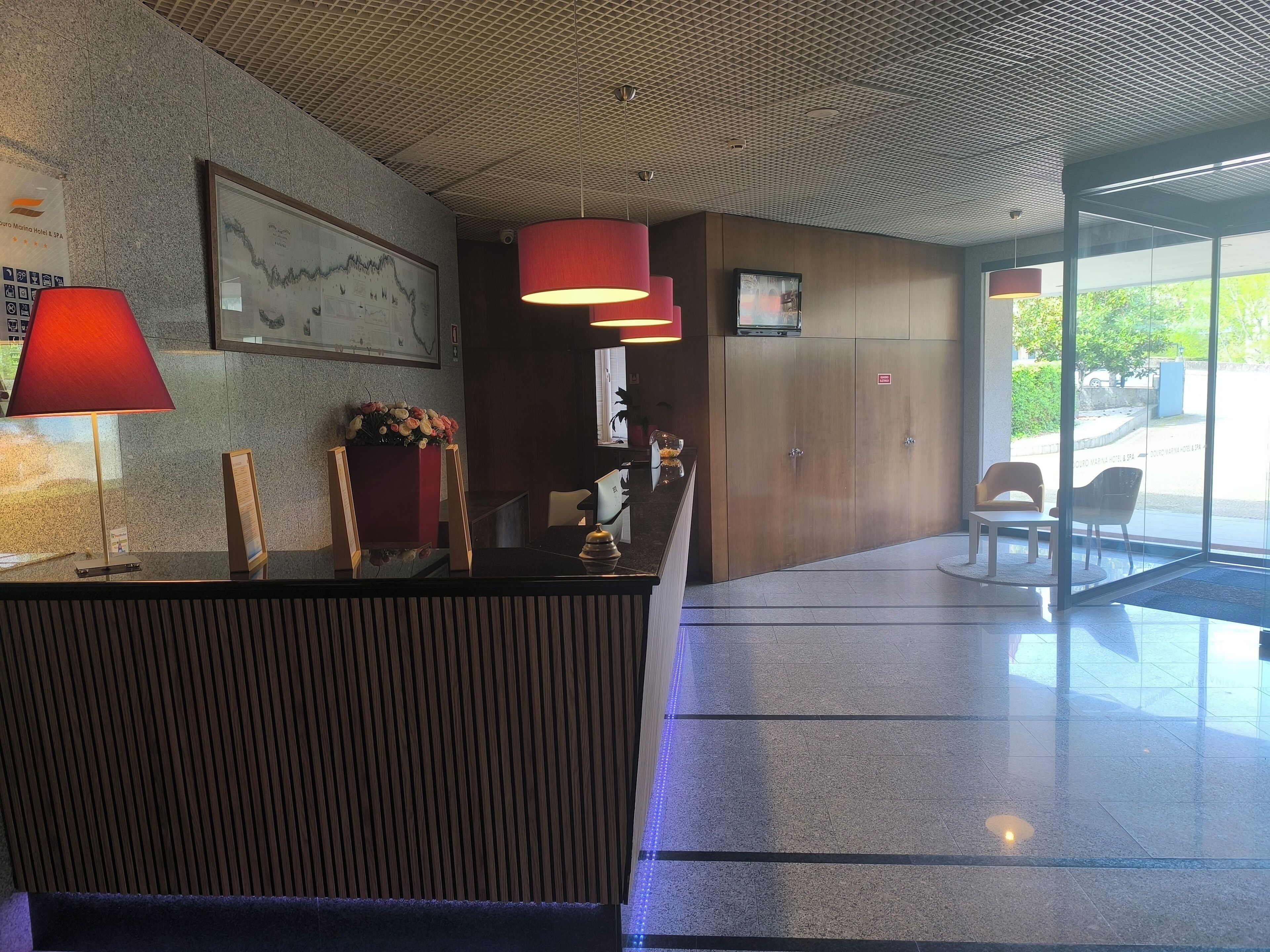 Un espace d’accueil d’hôtel comprenant un comptoir de réception, une lampe rouge, une carte encadrée et un coin salon avec des chaises et une table.