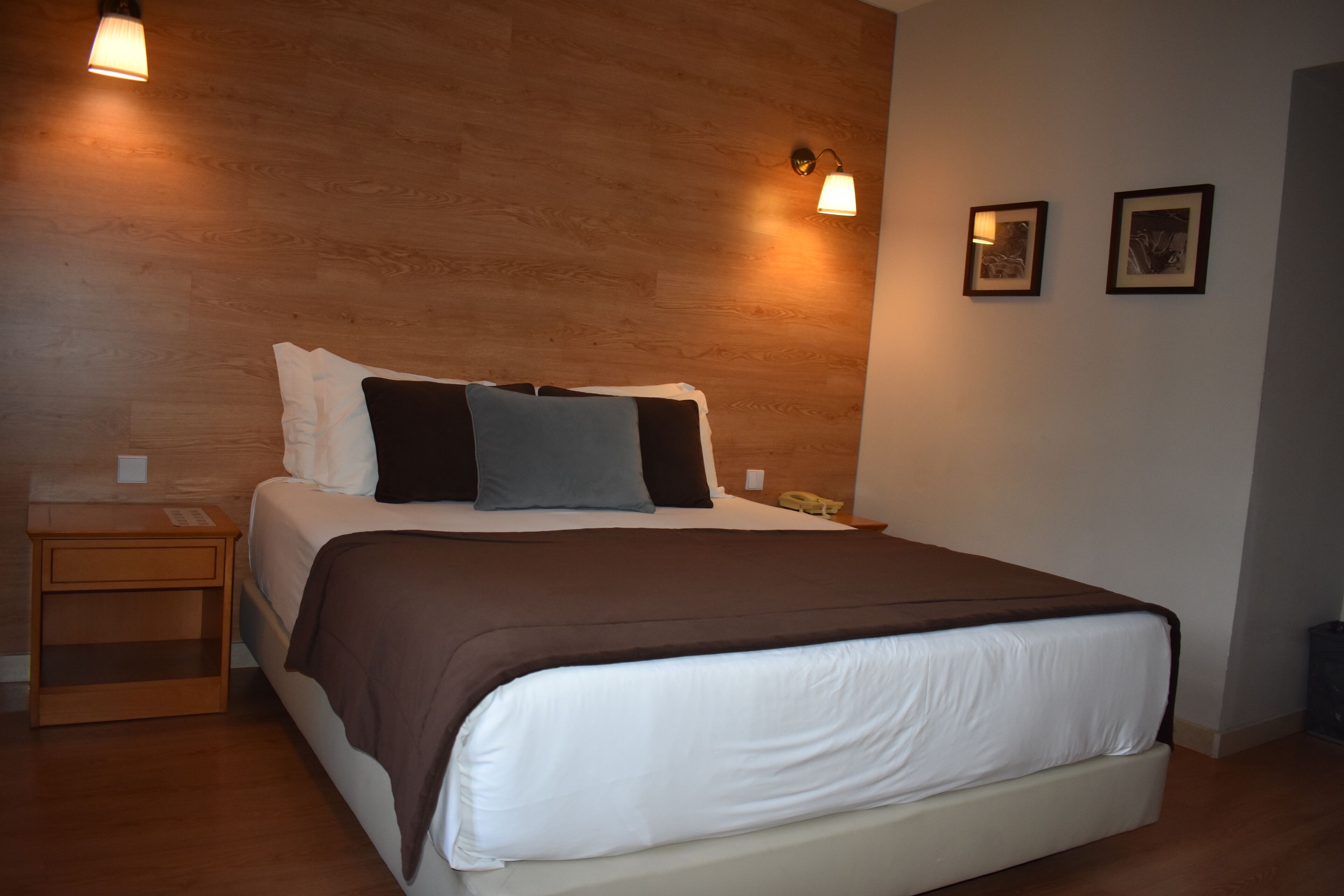 Un lit bien fait, recouvert d’une couvre-lit marron et blanche, avec deux lampes de chevet et deux tableaux encadrés accrochés au mur.