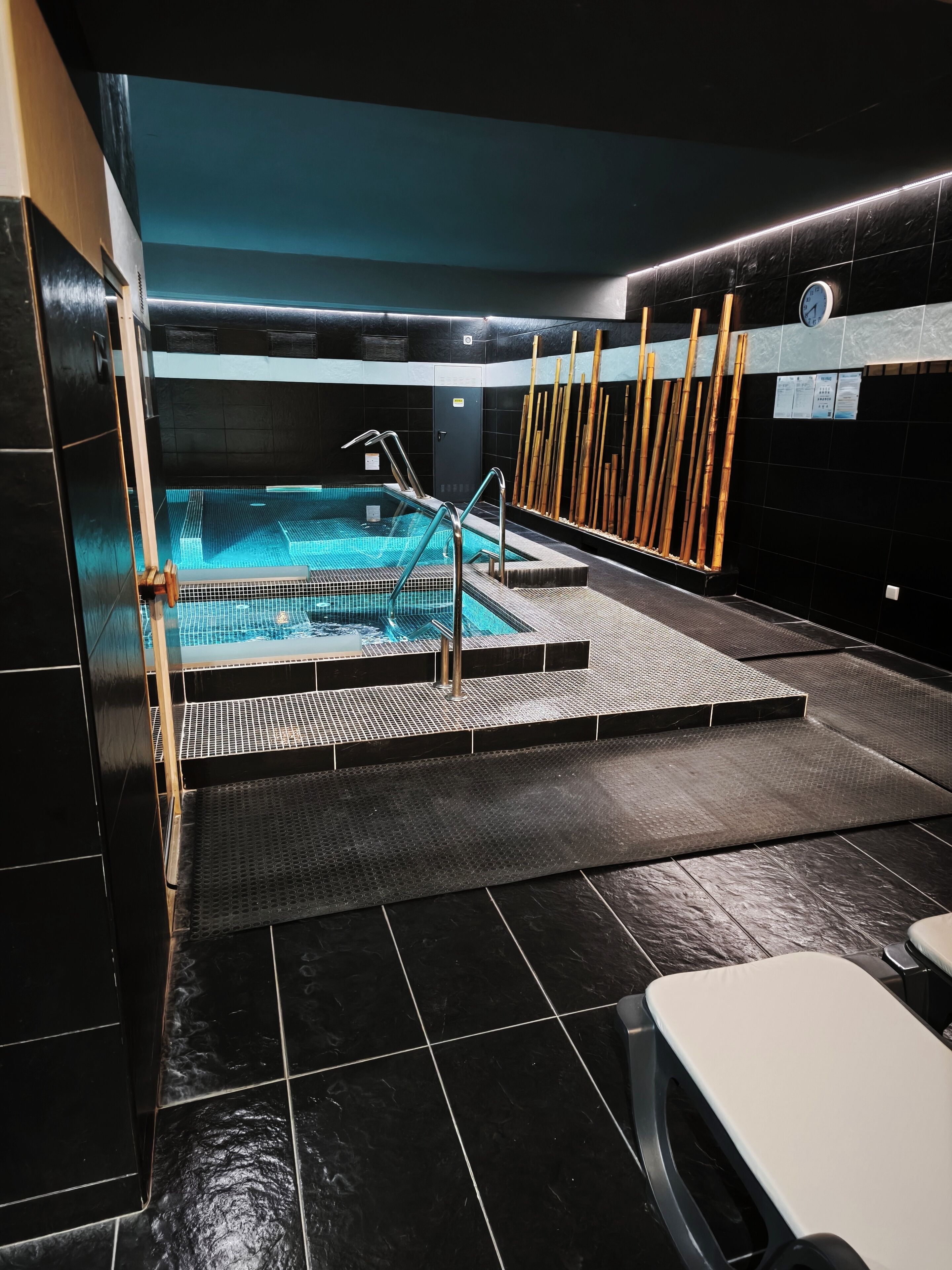 A modern indoor pool area with a clear water surface, metal steps leading into the pool, and bamboo sticks on the wall.