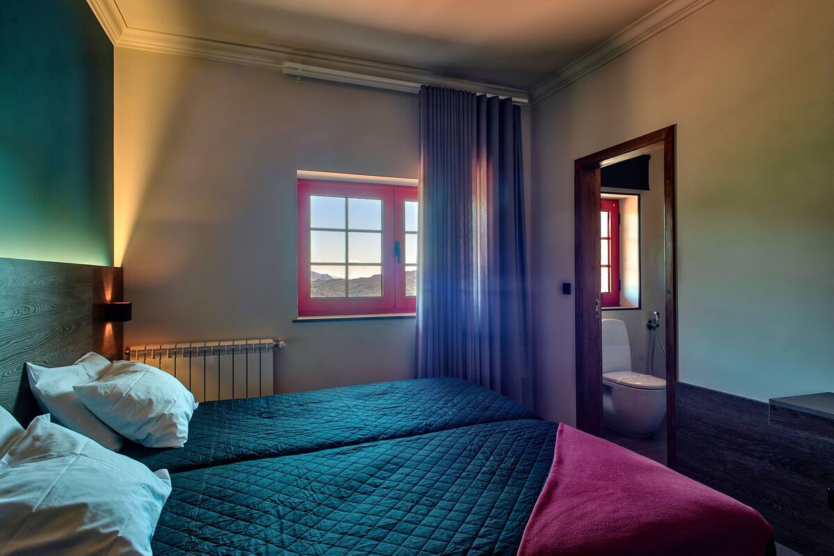A hotel room with two beds, a window with a view, a radiator, and a bathroom visible through an open door.