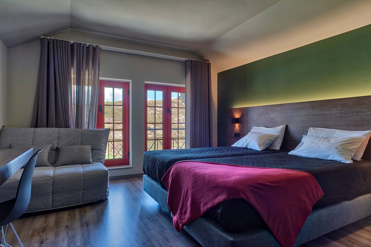 A bedroom with a bed, a sofa, and a chair. There is a window with curtains and a view outside.