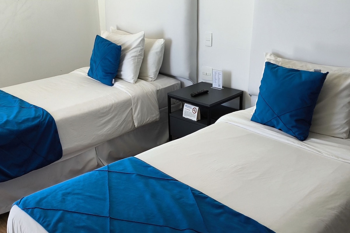 A hotel room with two single beds, each with a blue and white duvet, a small black nightstand with a remote control, and a sign indicating no smoking.