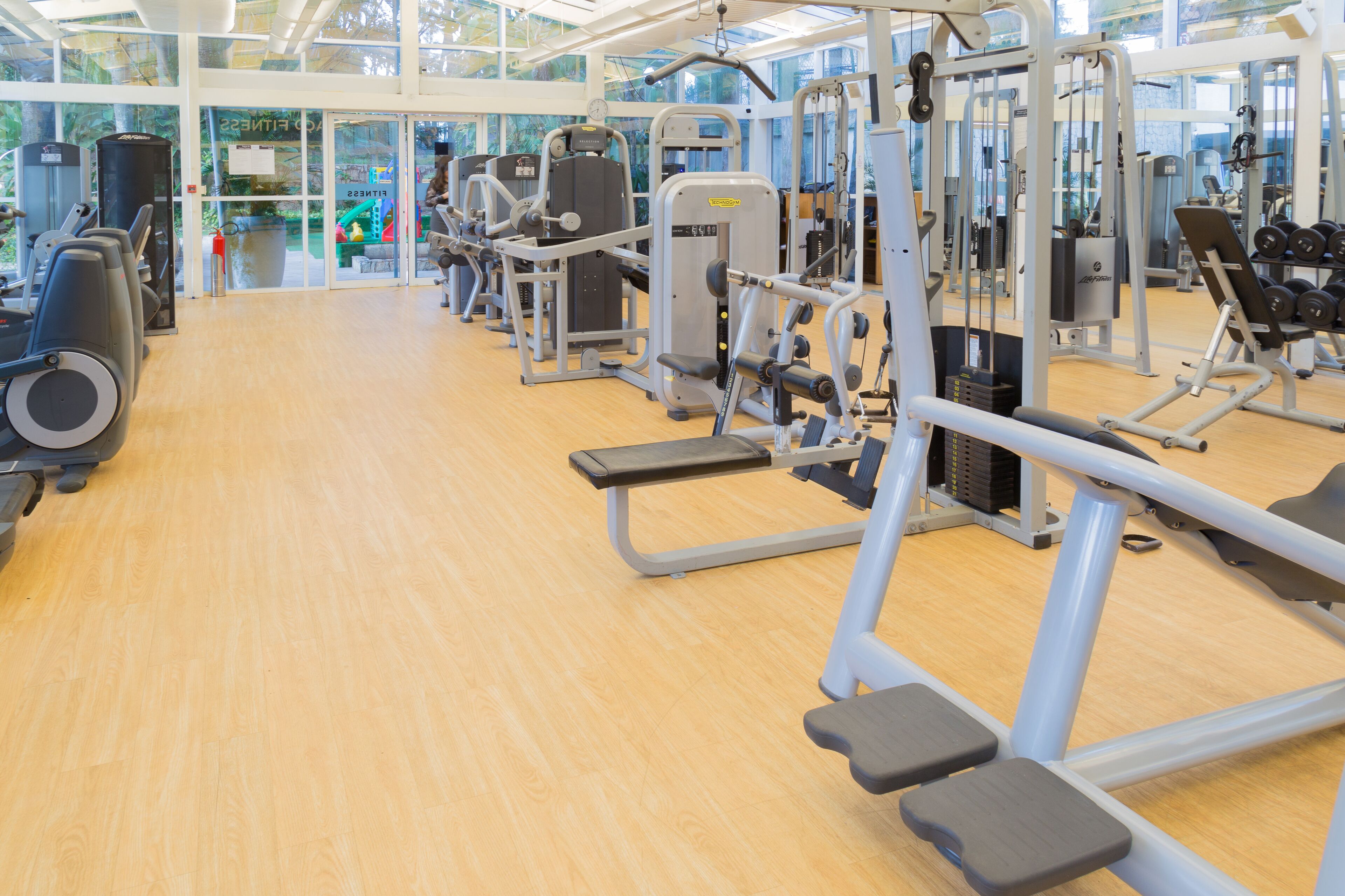 Un gimnasio con diversas máquinas de ejercicio, suelo de madera y amplias ventanas que dejan entrar luz natural.