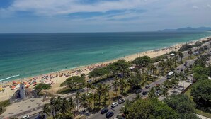 A beach with a road running parallel to it, lined with palm trees and parked cars. - Wyndham Rio Barra - Oficial (Rio de Janeiro)