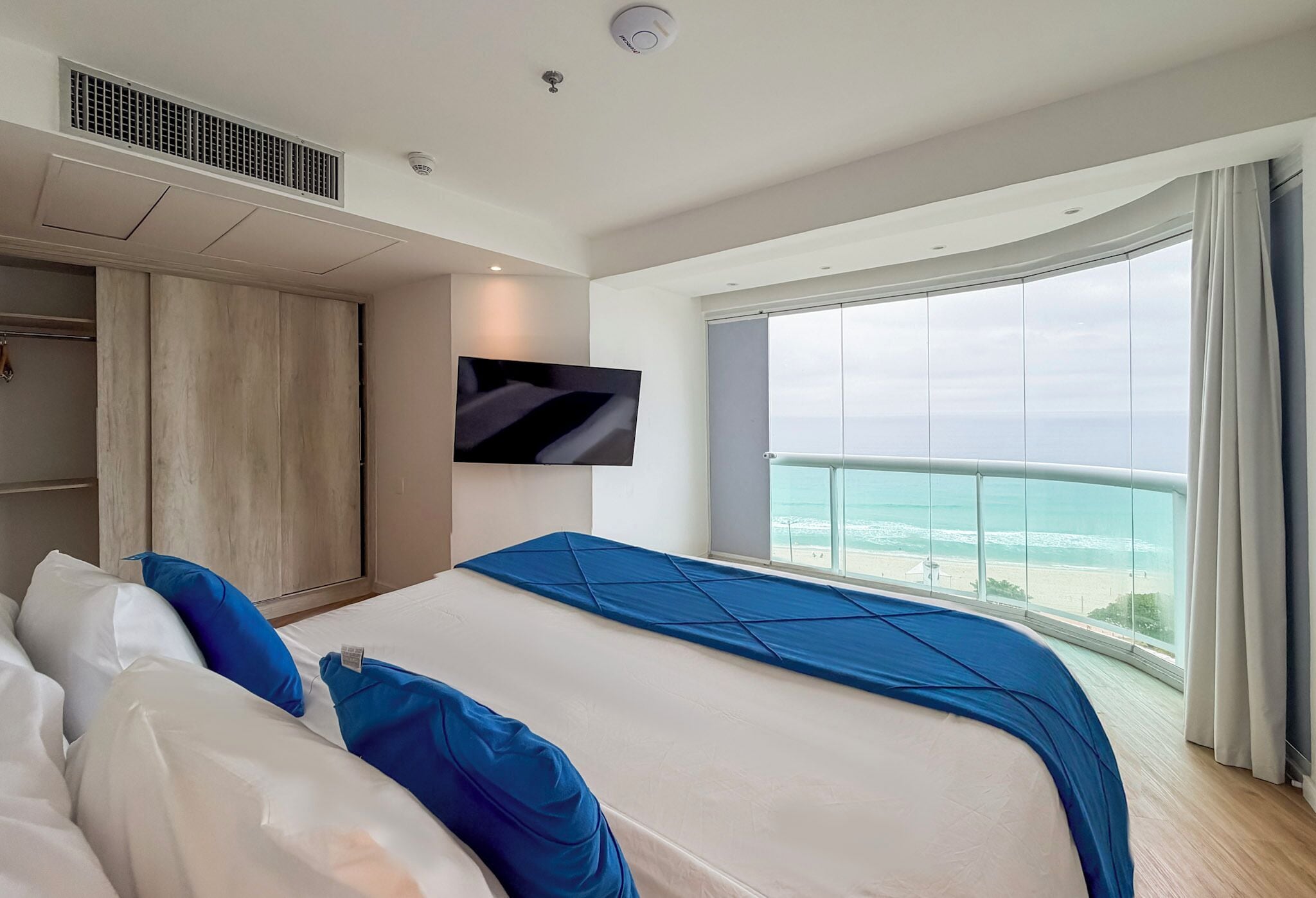 A modern hotel room with a large bed, a TV mounted on the wall, and a balcony with a view of the sea.