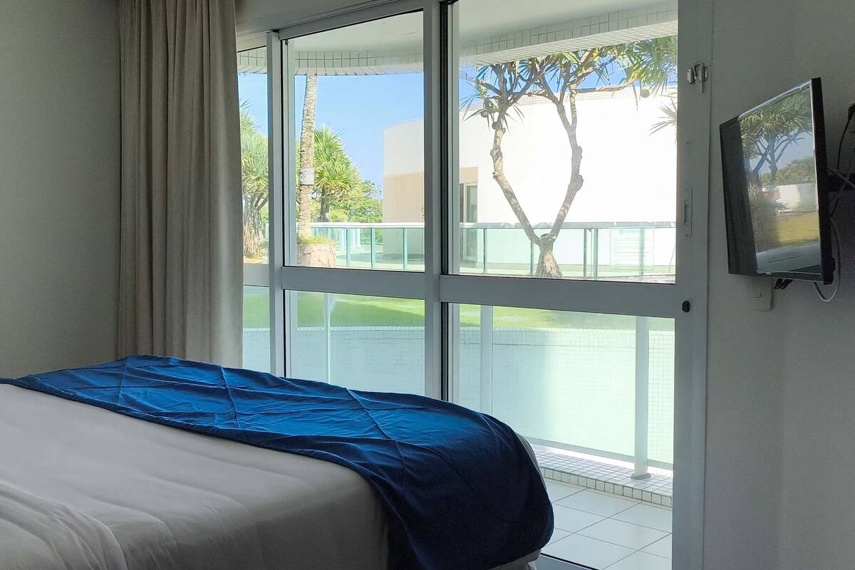 Ein Hotelzimmer mit einem Bett, einem an der Wand montierten Fernseher und einer Glasschiebetür, die zu einem Außenbereich mit Blick auf Palmen und ein Gebäude führt.