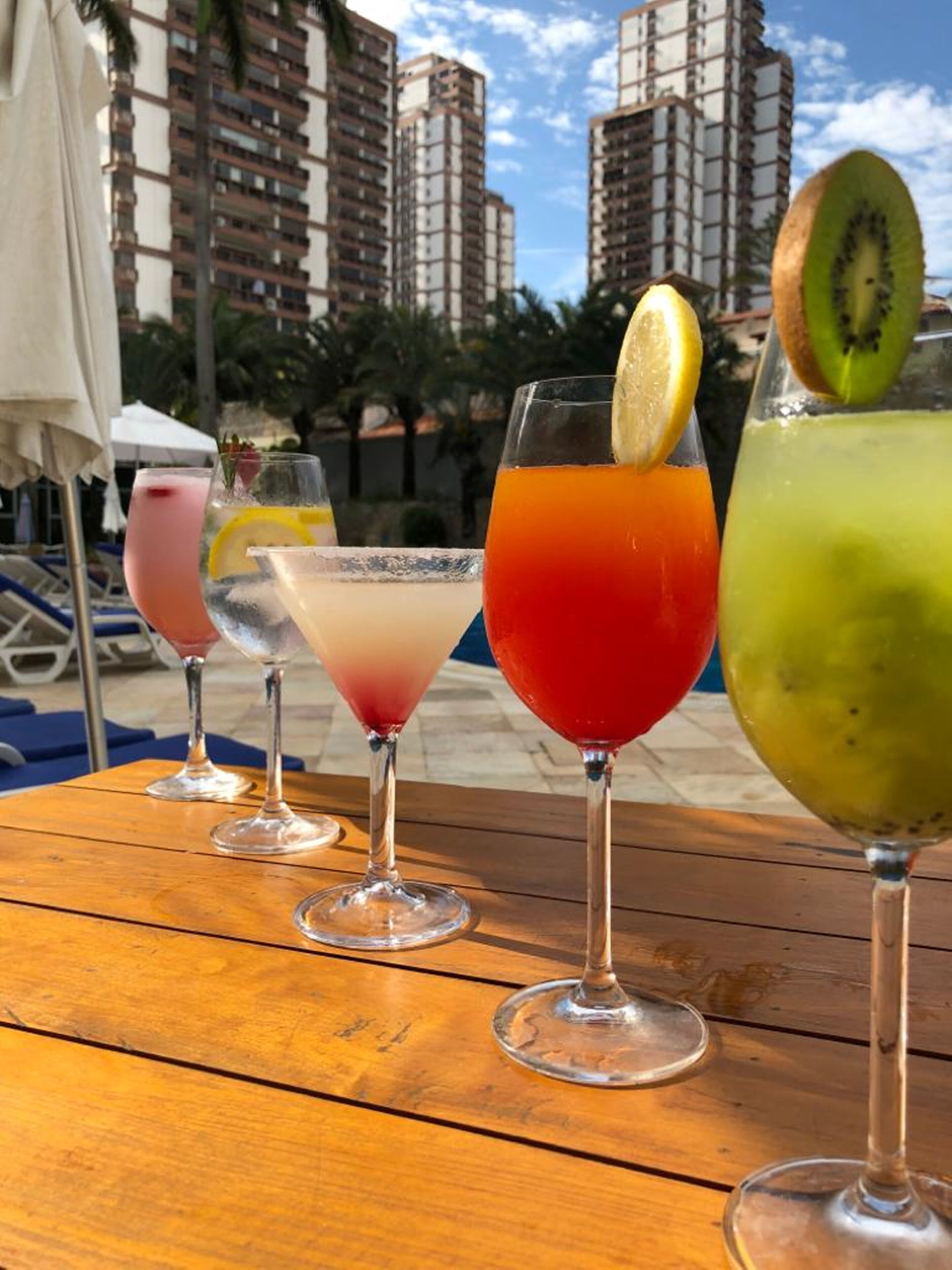 A row of colorful cocktails on a wooden table with a poolside view in the background.