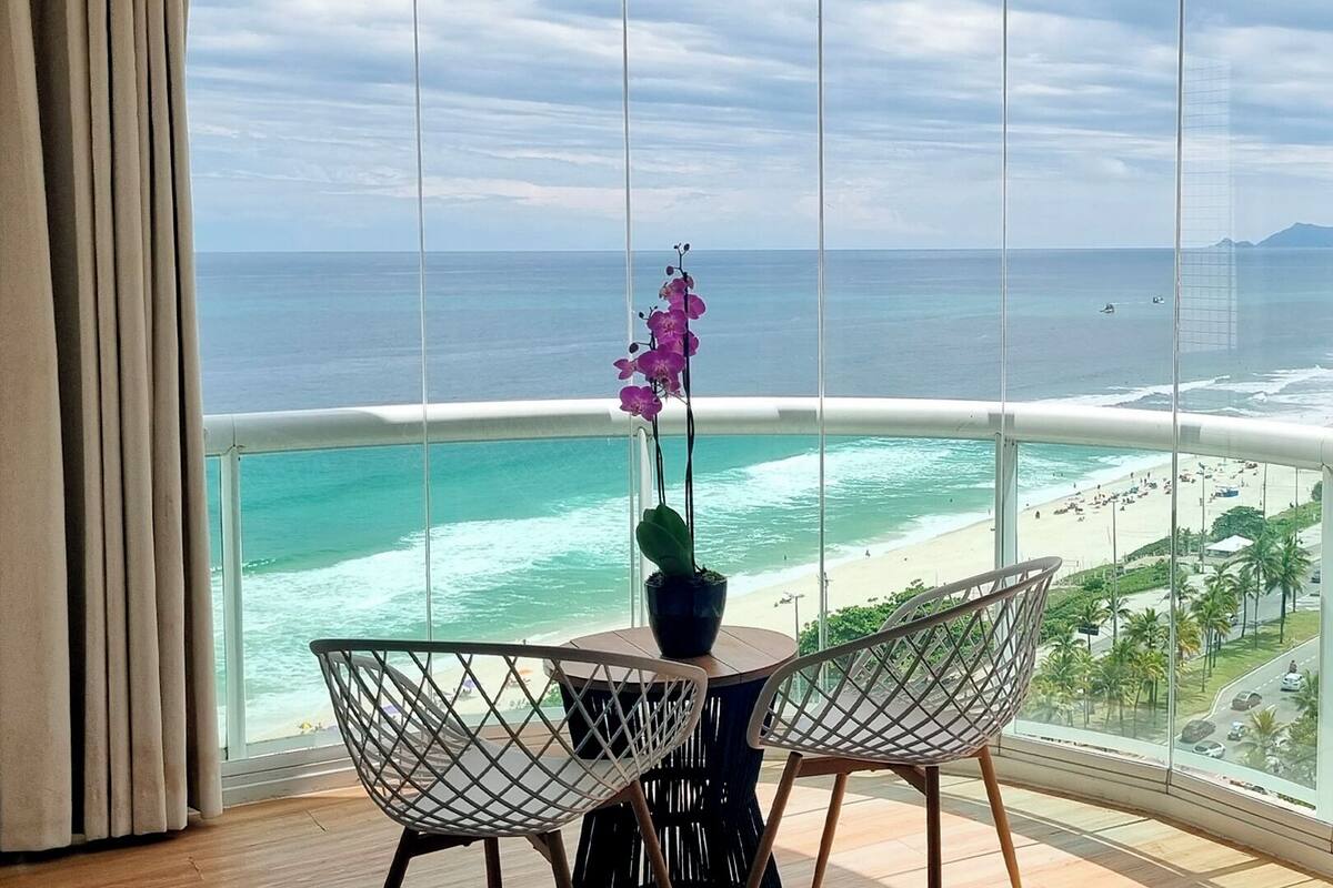Ein Balkon mit Blick auf den Ozean und den Strand, zwei Stühle und ein Tisch mit einer Topforchidee.