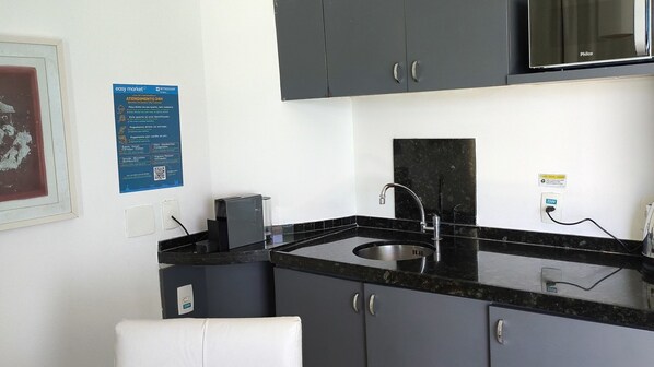 A small kitchenette with a sink, cabinets, and a countertop. There is a poster on the wall and a power outlet with a 220V label.