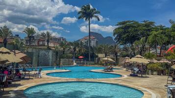 A pool area with multiple circular pools, lounge chairs, umbrellas, and palm trees.