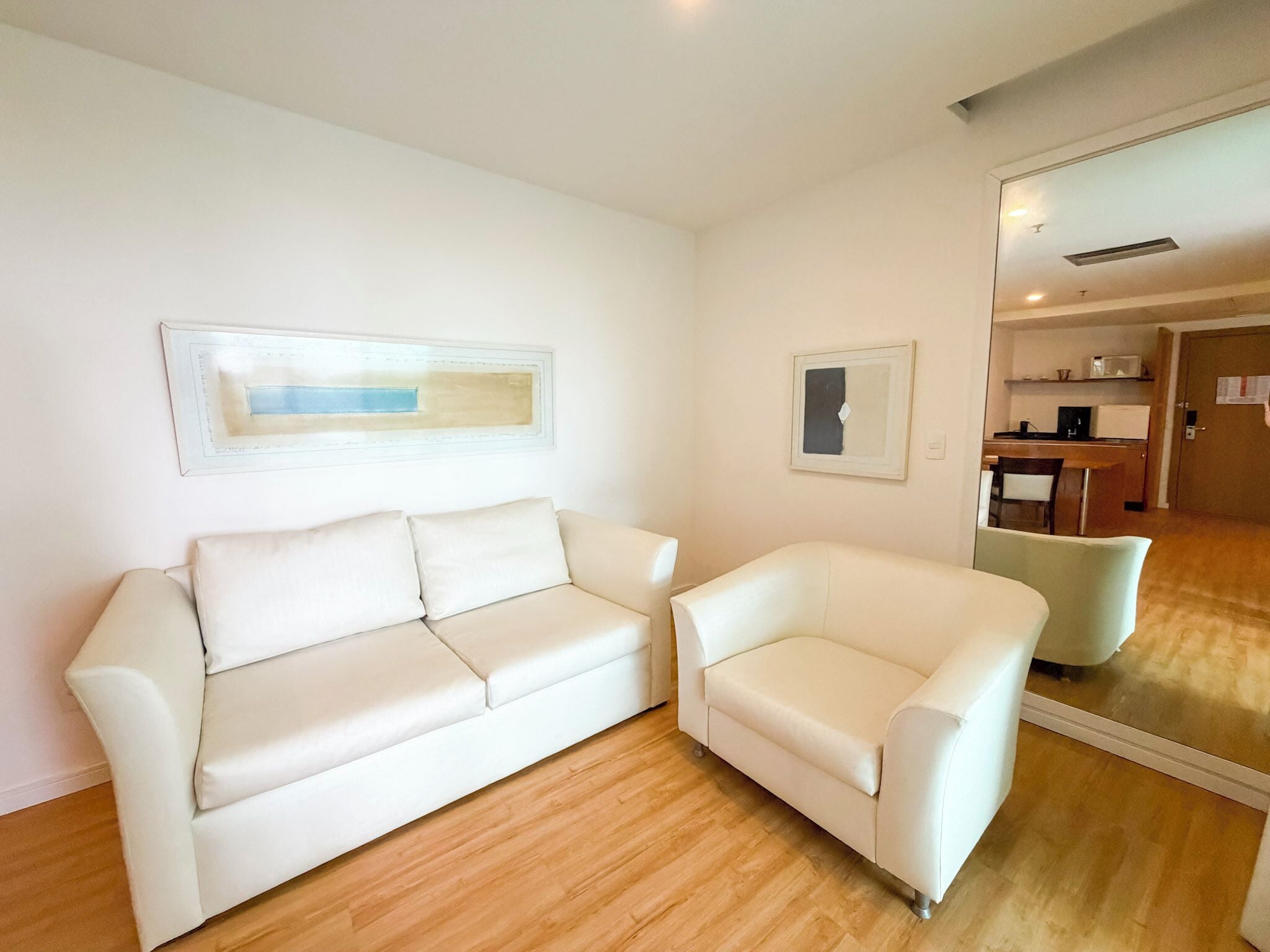 A modern living room with a white sofa and armchair, wooden flooring, and a kitchen area visible through an open door.