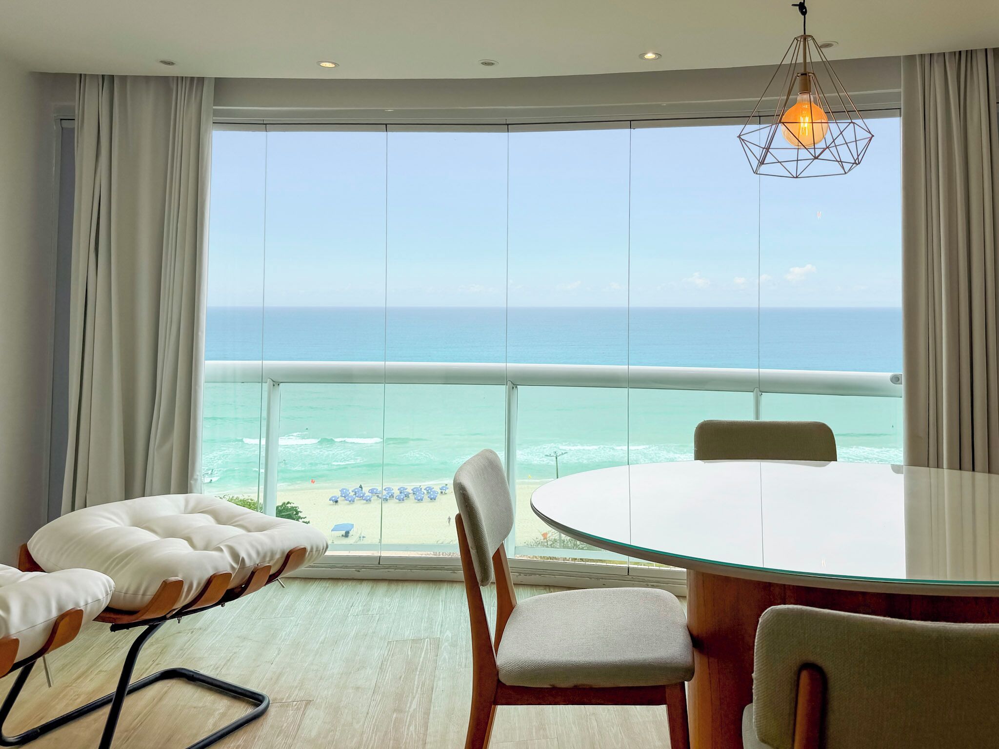 Una habitación con un ventanal grande con vistas a una playa, una mesa redonda de vidrio, dos sillas y una lámpara colgante.