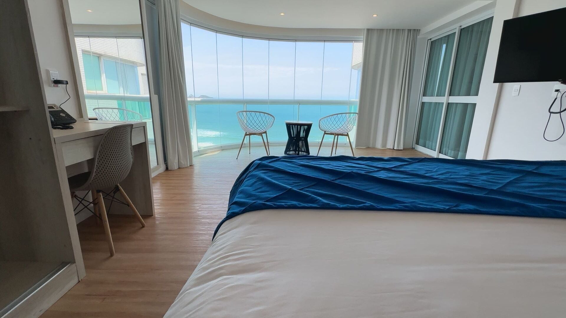 Quarto de hotel com cama, escrivaninha, duas cadeiras e uma grande janela com vista para o mar.