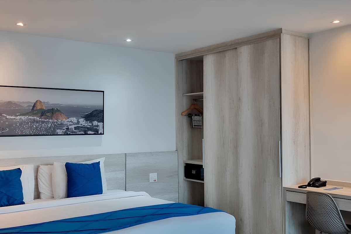 Ein Hotelzimmer mit Bett, Schreibtisch, Stuhl, Kleiderschrank und einem an der Wand montierten Fernseher, der eine Stadtansicht zeigt.