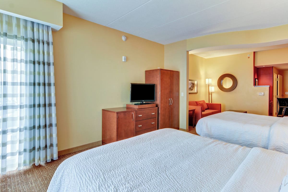 Habitación de hotel con dos camas, un televisor sobre un mueble de madera, un escritorio con una silla y un baño visible a través de una puerta abierta.