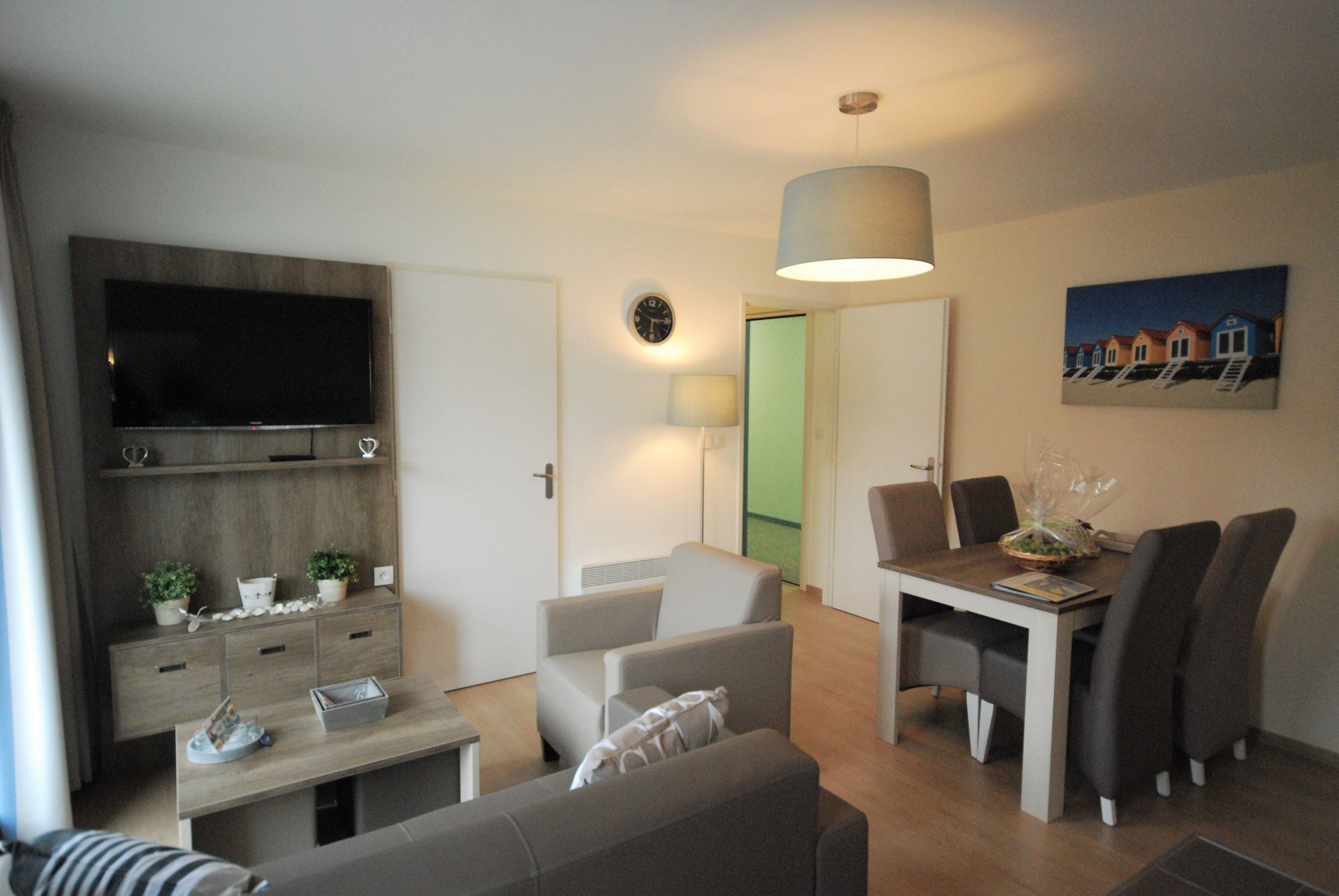 A modern living room with a flat-screen TV, a dining table with chairs, a sofa, a coffee table, and a lamp.