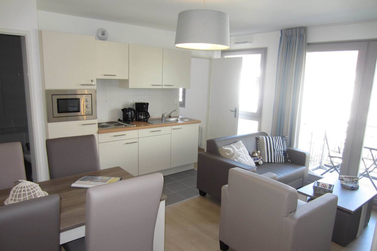 Cocina moderna equipada con microondas, cafetera y lavaplatos. Cuenta con un comedor con mesa y sillas, además de una sala de estar con sofá y cojines.