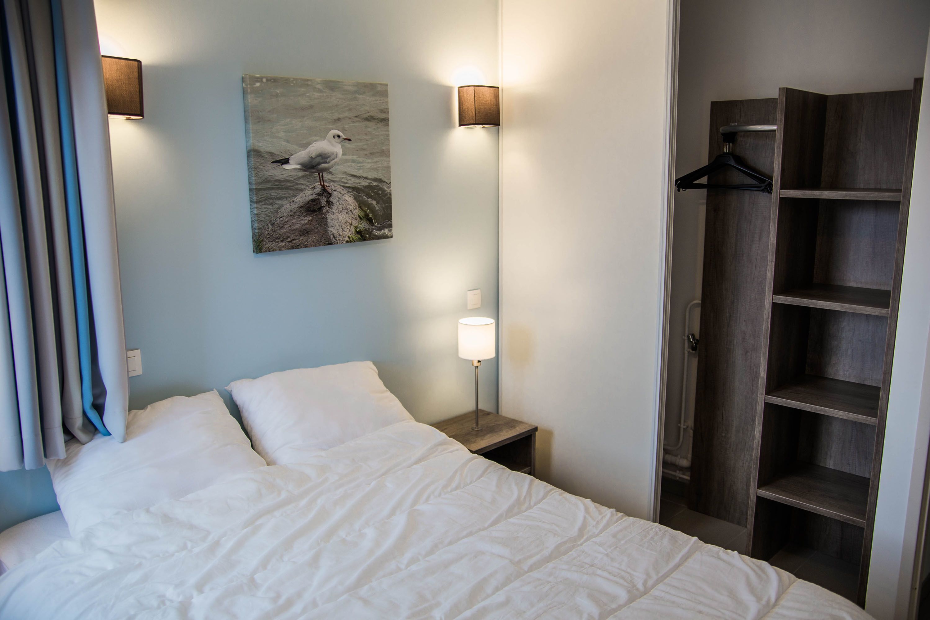Een netjes opgemaakt bed met witte lakens, een aan de muur hangend schilderij van een meeuw, en een houten garderobe met kledingroede.