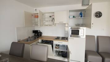 A compact kitchen with a microwave, stove, and coffee maker. There are cabinets with dishes and a clock on the wall.