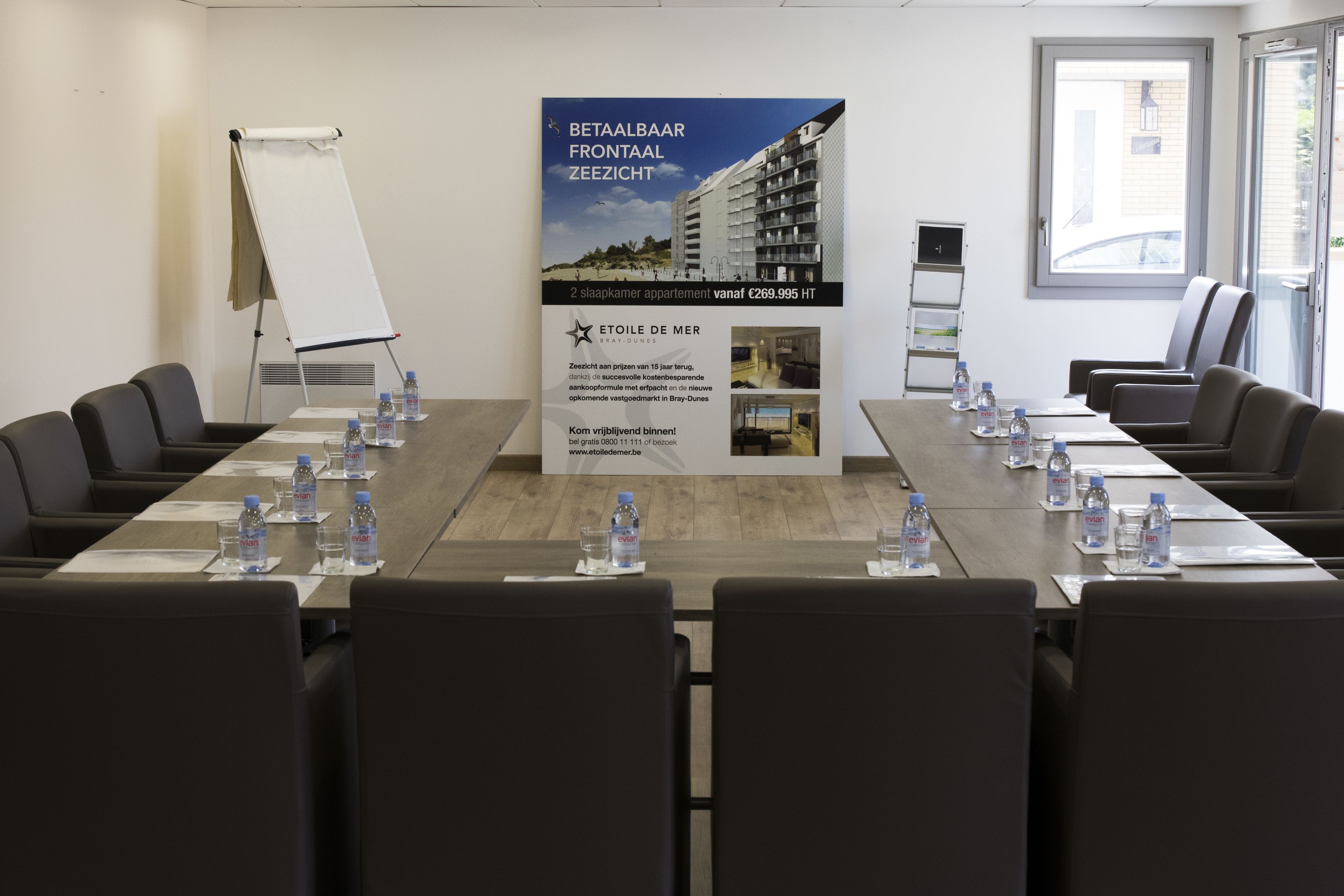 A conference room with a long table, chairs, water bottles, and a poster advertising a hotel.