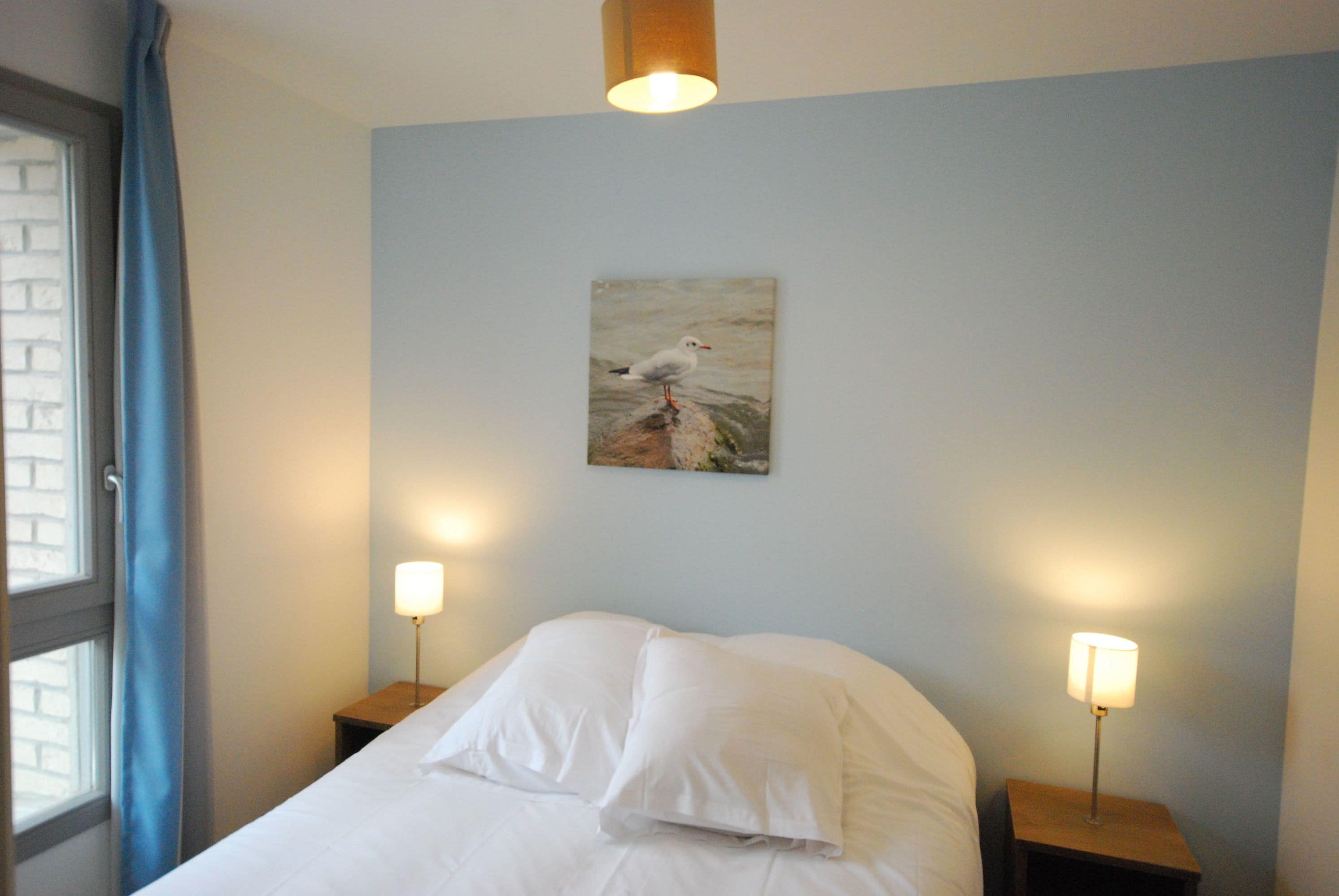 Habitación de hotel con una cama, dos lámparas de noche, una ventana con cortinas y un cuadro en la pared.