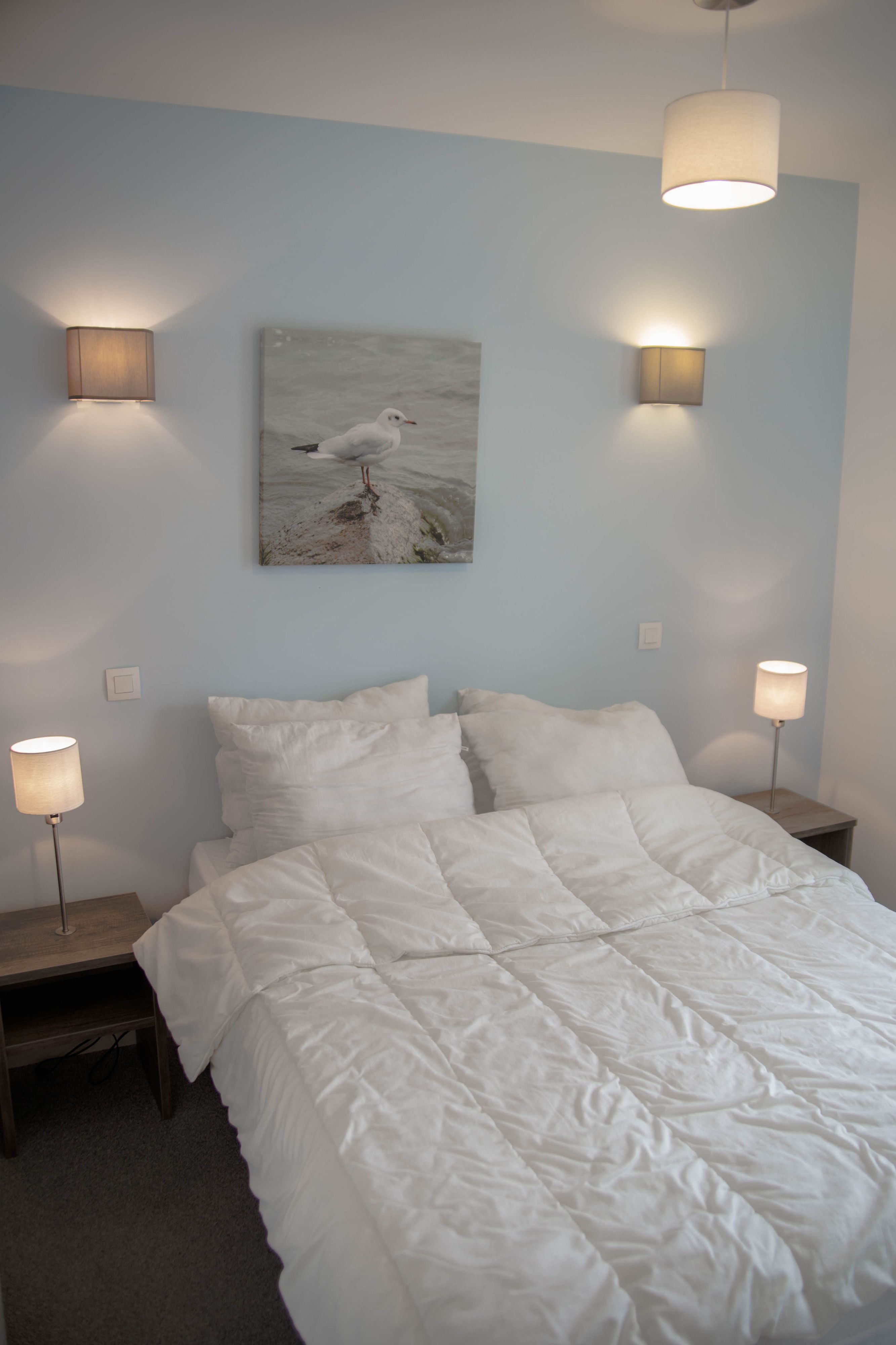 A neatly made bed with white bedding, two bedside lamps, and a wall-mounted painting of a seagull.