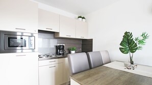 A modern kitchen with white cabinetry, a built-in microwave, and a coffee maker. There is a dining table with two chairs and a large plant on the table. - Evancy Bray-Dunes (Bray-Dunes)