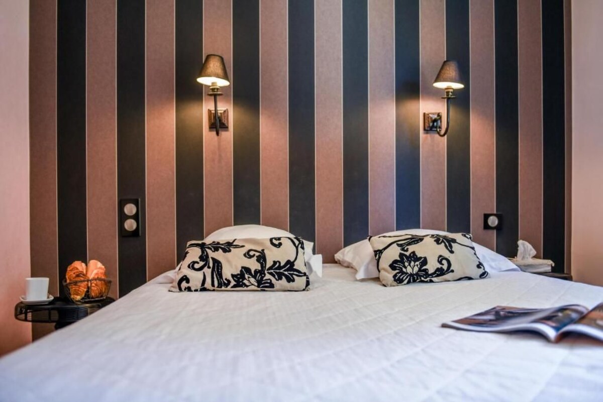A hotel room with a striped wall, two bedside lamps, a bed with floral pillows, a nightstand with a cup and croissants, and a magazine on the bed.