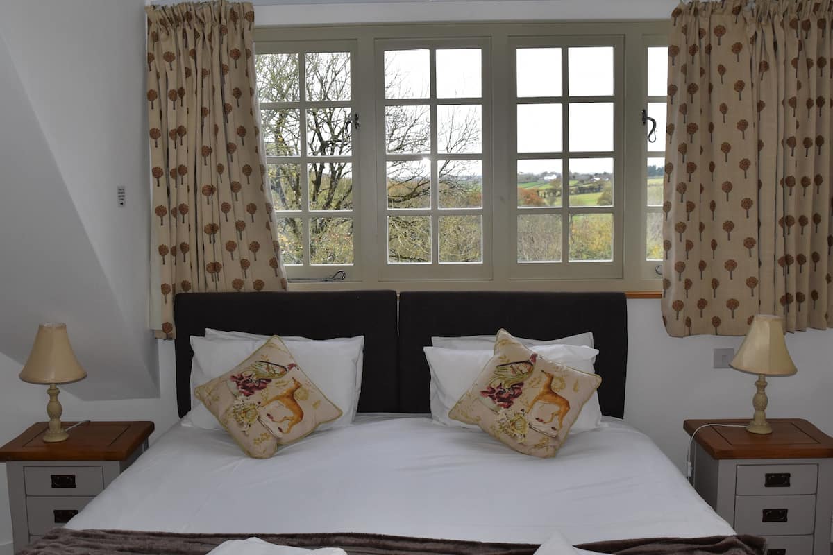 Quarto com cama grande, duas mesas de cabeceira com luminárias, janela com cortinas e vista para árvores e campos.