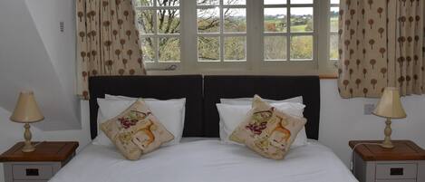 A bedroom with a large bed, two bedside tables with lamps, a window with curtains, and a view of trees and fields.